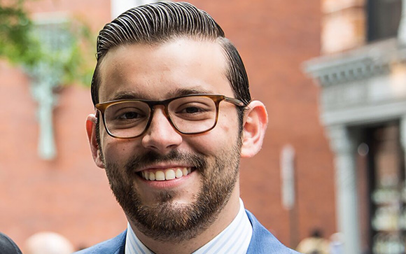 A smiling man with dark hair, glasses, and a beard wearing a blue suit jacket.