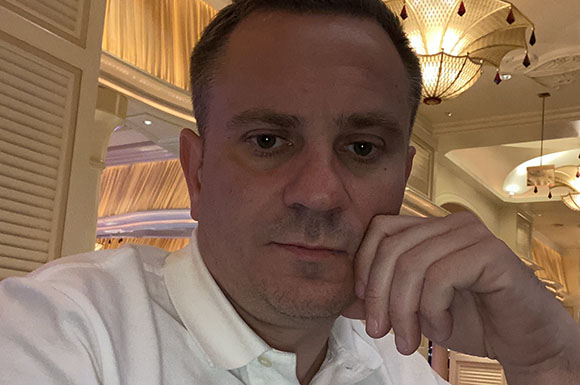 A man in a white polo shirt rests his chin on his hand in a room with ornate lighting.