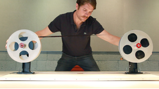A man in a black polo shirt adjusts a strip of film between two large metal reels.