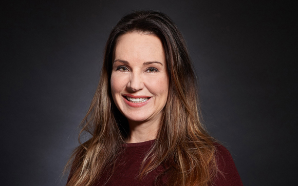 A smiling woman with long brown hair wearing a maroon top against a dark background.