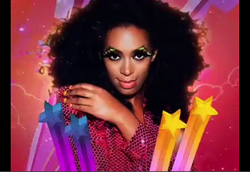 A woman with large dark hair and colorful eye makeup poses against a pink background with stars.