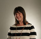 A woman with brown hair and bangs smiling, wearing a black and white striped shirt and beaded necklaces.