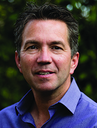 A middle-aged man with short dark hair and a blue button-down shirt smiling against a leafy background.