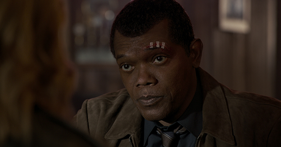 A man with a small bandage on his forehead looks toward a person in a dimly lit room.