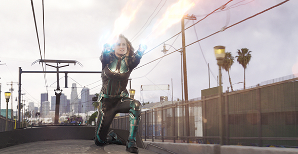 A woman in a dark superhero suit kneels on a train track, shooting blue energy from her hands.