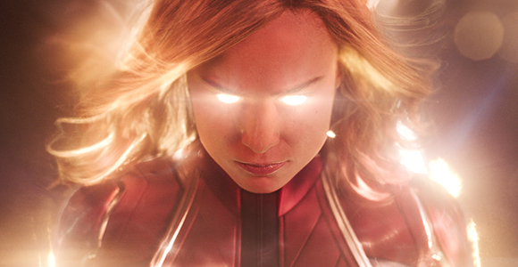 A woman in a red suit with glowing white eyes and flowing blonde hair surrounded by light.