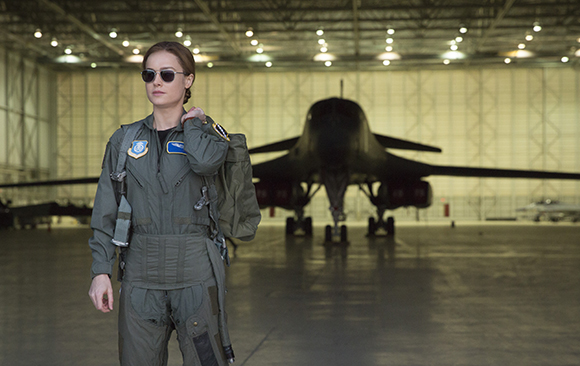 A woman in a green flight suit and sunglasses stands in a hangar before a large black bomber jet.