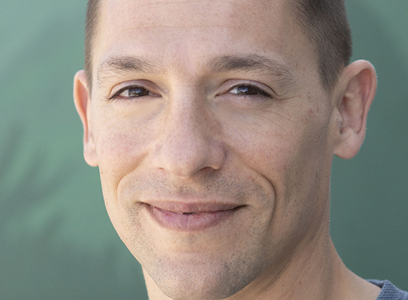 Close-up portrait of a man with short hair and brown eyes smiling against a green background.