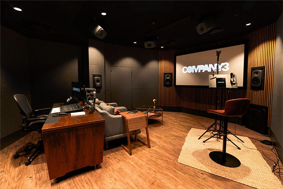 A dimly lit recording studio featuring a wooden desk, a grey sofa, and a large screen displaying COMPANY 3.