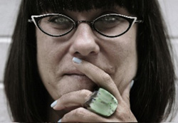 Close-up of a person with dark hair and cat-eye glasses, resting a hand with a large green ring on their chin.