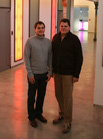 Two men standing side-by-side in a brightly lit hallway with colorful light panels.
