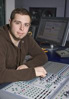 A man in a brown hoodie sits at a desk with a mixing console and computer monitor in a studio.