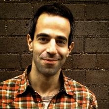 A man with dark hair and a slight smile wearing an orange and brown plaid shirt against a dark brick wall.