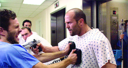 A man in a patterned hospital gown holds a handgun toward a man in blue scrubs in a hallway.