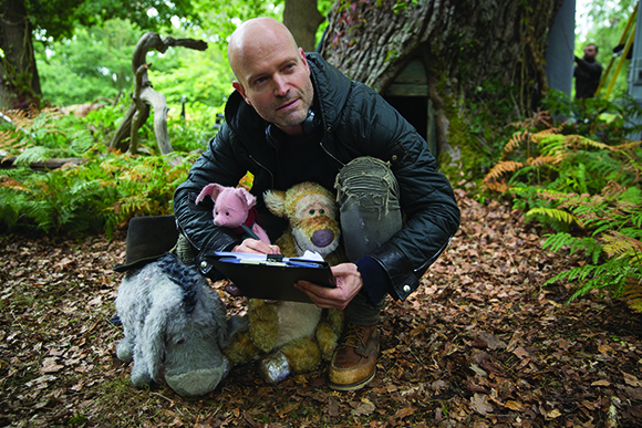 A bald man in a black jacket crouches in a forest among ferns and stuffed animal puppets.