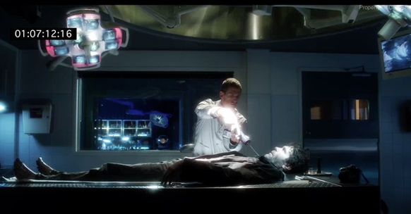 A person in a white lab coat uses a glowing tool on a patient lying on a table in a dark medical room.