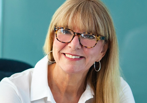 A smiling woman with blonde hair, glasses, and hoop earrings wearing a white shirt.