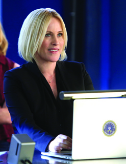 A blonde woman in a black blazer sits at a desk using a laptop with a circular emblem on the lid.