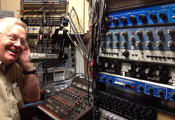 A man smiles next to a microphone and stacks of professional audio mixing and processing equipment.