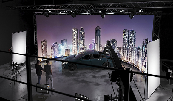 A car sits on a studio set in front of a large purple-hued city skyline backdrop with crew members nearby.