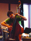 A person in a green shirt playing a large wooden double bass in an indoor setting.
