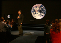 A man in a suit stands on a stage next to a large projection of Earth, facing an audience in a dark room.