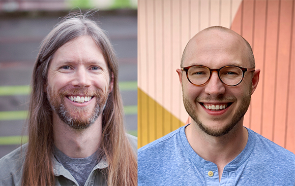 Two smiling men; one with long hair and a beard, the other bald with glasses and a beard.