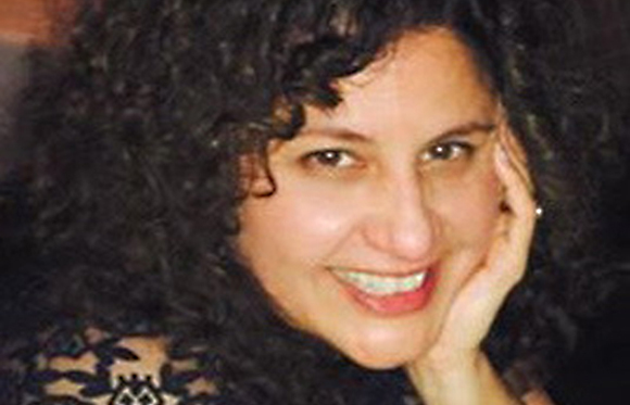 A woman with dark curly hair smiling with her hand resting on her cheek.
