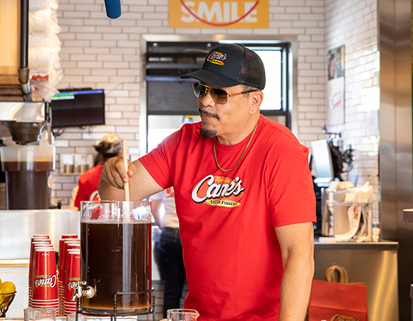 A man wearing a red Cane's t-shirt and a black cap stirs a large dispenser of iced tea in a restaurant.