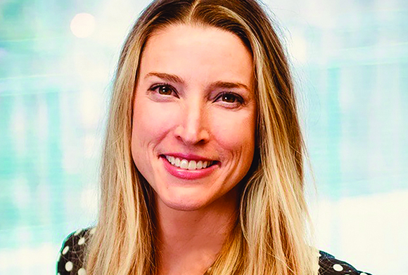 A smiling woman with long blonde hair looking toward the camera against a blurred light blue background.