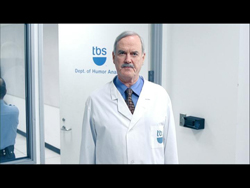 A man with a mustache wearing a white lab coat and tie stands in a clinical hallway.