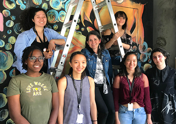Seven young women pose together in front of a large, colorful mural with a silver ladder in the center.