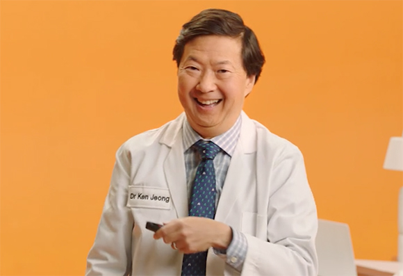 A smiling man wearing a white lab coat and patterned blue tie against a solid orange background.