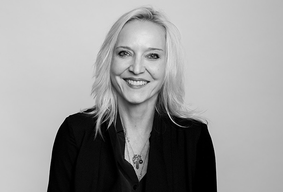 A smiling blonde woman wearing a dark shirt and necklace, looking at the camera against a plain background.
