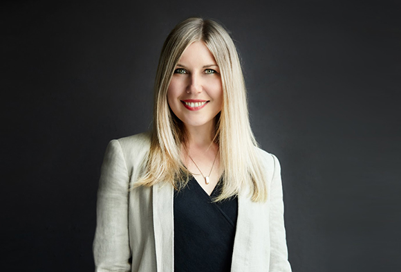 A smiling woman with long blonde hair, wearing a white blazer and black top, against a dark background.
