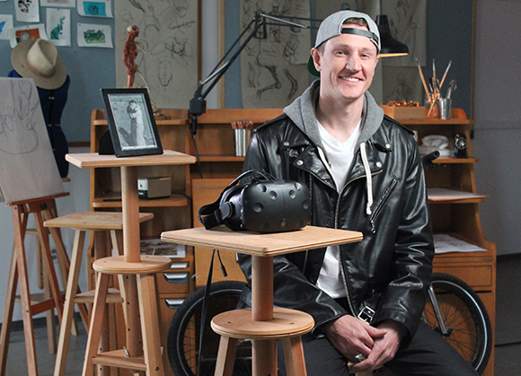 A smiling man in a black leather jacket sits in an art studio behind a VR headset on a wooden stand.