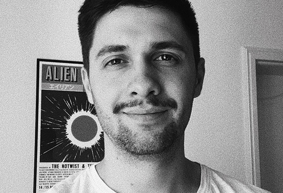Black and white portrait of a man with a mustache smiling in front of an Alien movie poster.