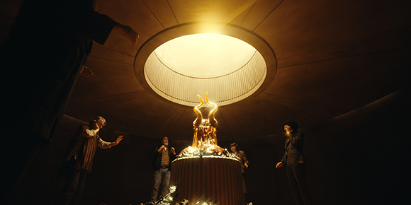 Four people stand in a dark, circular room around a glowing golden horned sculpture under a bright skylight.