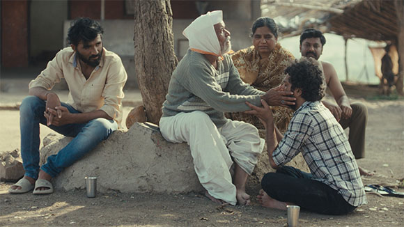 A group of five people sit together in a dusty outdoor area near a tree and a thatched structure.