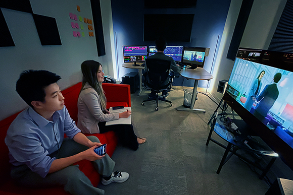 Two people sit on a red couch watching a large screen in a dark studio with multiple computer monitors.