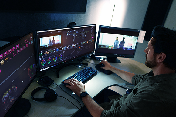 A man sits at a desk with three large monitors displaying video editing software and footage.