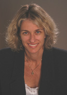 A smiling woman with shoulder-length blonde hair wearing a black blazer and a necklace.
