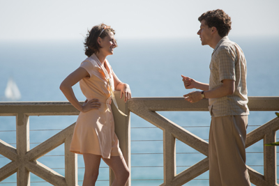 A man and a woman in vintage clothing talk on a balcony with a view of the sea.
