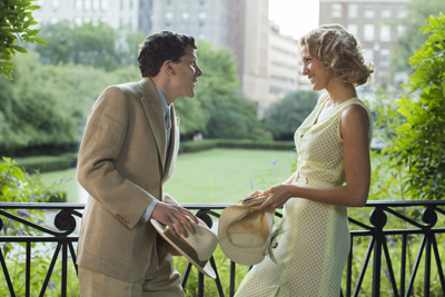 A man in a tan suit and a woman in a yellow dress talk near a black metal railing in a park.