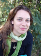 A woman with long brown hair and a green scarf smiles in front of green foliage.