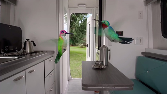 Two colorful hummingbirds flying through the interior of a white camper van near a table and kitchen.
