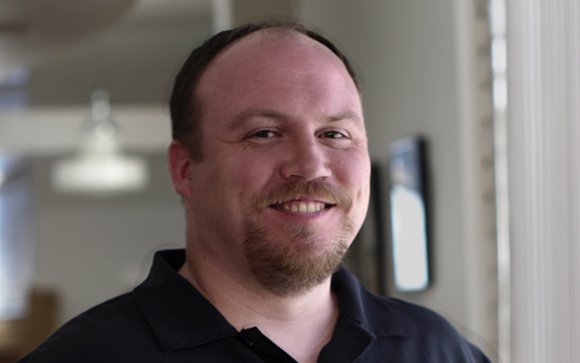 A smiling man with short brown hair and a goatee wearing a black polo shirt.