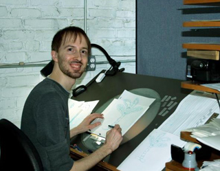 A man with short hair and a beard sits at a drafting desk covered in technical drawings.