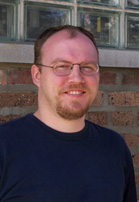A man with glasses and a goatee smiling in front of a brick wall and glass blocks.