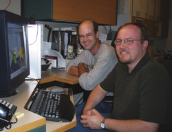 Two men smiling while sitting at a desk in front of a computer monitor.